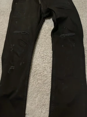 Svarta ripped jeans ICON2, stl 34 - Svarta jeans från ICON2 i storlek 34 med slitna detaljer vid knäna. Tillverkade i 98% bomull och 2% elastan för en bekväm stretchig passform. Perfekta för en edgy och avslappnad stil.