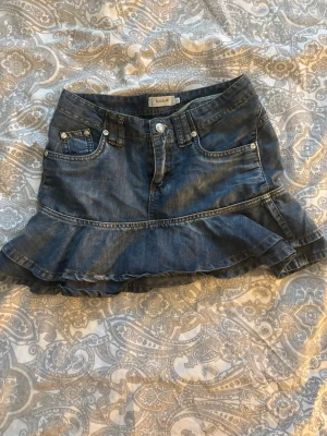 Jeanskjol med volang från Miss E-vie - Säljer en blå jeanskjol från Miss E-vie med volangkant nedtill och klassiska jeansdetaljer som fickor och bälteshällor. Kjolen har en kort modell och stängs med knapp och dragkedja framtill. Perfekt för dig som gillar denim och vill ha en söt twist.