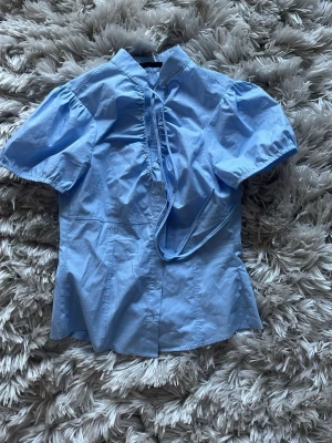 Ljusblå blus med puffärm och knyt - Säljer denna trendiga topp. Snygg ljusblå blus med puffiga korta ärmar och knytband vid halsen. Blusen har en rak passform och diskreta sömmar som ger en feminin siluett. Tillverkad i ett tunt, vävt material som känns lätt och fräscht.