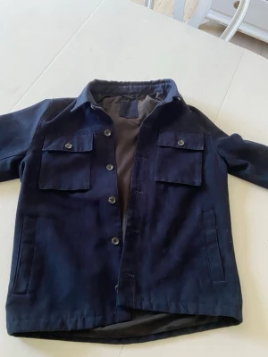 Mörkblå overshirt från Next - Stilren mörkblå overshirt från Next med två bröstfickor med lock och knapp, samt två sidofickor. Klassisk krage och knäppning framtill. Materialet är mjukt och har en lätt fodrad insida för extra komfort. Perfekt lager på lager-plagg.