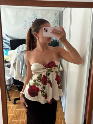 Blommig axelbandslös topp med rosett - Säljer en söt axelbandslös topp i vitt med stora röda rosor och gröna blad. Toppen har en rosett framtill och ett flowy fall. Perfekt för sommarens alla tillfällen och ger en romantisk vibe. Från Urban outfitters 💕