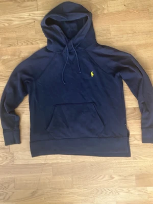 Mörkblå hoodie från Polo Ralph Lauren - Mörkblå hoodie från Polo Ralph Lauren med klassisk gul logga på bröstet. Hoodien har huva med dragsko, stor magficka och ribbade muddar vid ärmar och nederkant. Tillverkad i mjukt bomullsmaterial, perfekt för en avslappnad och stilren look.