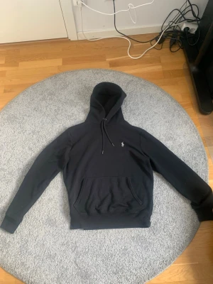 Svart ralph lauren hoodie - Säljer en svart hoodie med huva och dragsko samt en klassisk känguruficka framtill. På bröstet finns en liten broderad logga i vitt. Hoodien har ribbade muddar vid ärmar och nederkant. Perfekt för en chill och stilren look.