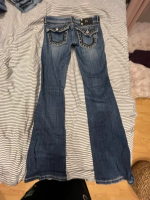Blå bootcut jeans med detaljer - Säljer ett par blå bootcut jeans med snygga slitningar och dekorativa sömmar på bakfickorna. Jeansen har klassisk femficksmodell och är tillverkade i denim. Perfekta för dig som gillar en retro och avslappnad stil.