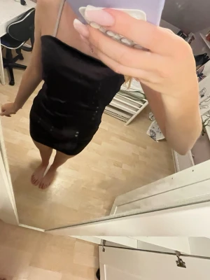 Svart kort klänning med smala band - Säljer en svart, tight kort klänning med smala axelband. Klänningen har en enkel och stilren design och är gjord i ett glansigt material som ger en snygg look. Perfekt för dig som gillar minimalistisk stil.