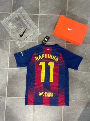 Barcelona fotbollströja Raphinha 11 - Säljer en FC Barcelona fotbollströja med Raphinha och nummer 11 på ryggen. Tröjan har blå och vinröda vertikala ränder, klubbmärke och Spotify-logga på bröstet samt UNHCR-logga nedtill. Tillverkad i lätt och ventilerande material, perfekt för fotboll.