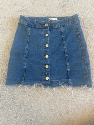 Blå jeanskjol med knappar framtill - Snygg blå jeanskjol med rå kant och knappar längs hela framsidan. Kjolen har en klassisk A-linjeform och är perfekt för dig som gillar en avslappnad men trendig stil. Säljer för att den inte kommer till användning 