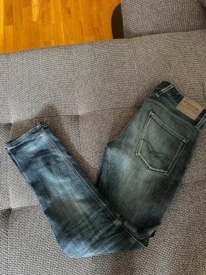 Replay blå slim fit jeans - Snygga blå slim fit jeans från Replay med klassisk femficksdesign och diskreta slitningar. Jeansen har en mörk tvätt med ljusare partier och Replay-logga på bakfickan samt läderpatch i midjan. Perfekta för en trendig och avslappnad look.