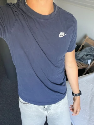 Mörkblå Nike t-shirt i bomull - Säljer en mörkblå t-shirt från Nike med klassisk vit logga på bröstet. T-shirten är tillverkad i mjuk bomull och har en normal passform med korta ärmar och rund hals. Perfekt för en avslappnad och sportig stil.