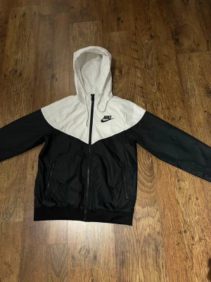 Svartvit Windbreaker Nike - Snygg svart och vit vindjacka från Nike med dragkedja framtill, huva och två fickor med dragkedja. Jackan har elastiska muddar vid ärmslut och nederkant samt klassisk Nike-logga på bröstet. Perfekt för en sportig och avslappnad stil. De kommer med free stickers. Pris är inte helt klart