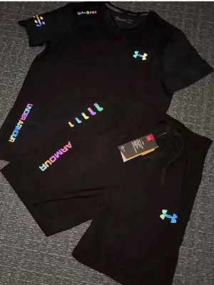 Svart träningsset från Under Armour - Snyggt svart träningsset med t-shirt och shorts från Under Armour. Setet har färgglada loggor och detaljer i regnbågsfärger. Perfekt för gymmet eller löprundan, tillverkat i ett lätt och snabbtorkande material.