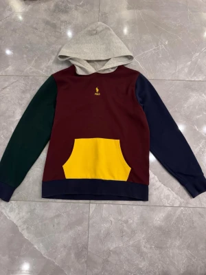 Flerfärgad hoodie från Polo Ralph Lauren - Snygg hoodie med grå huva, vinröd kropp, mörkgrön ärm, marinblå ärm och nederkant samt gul magficka. Liten gul Polo-logga på bröstet. Perfekt för dig som gillar färgblockad stil och vill sticka ut lite extra.