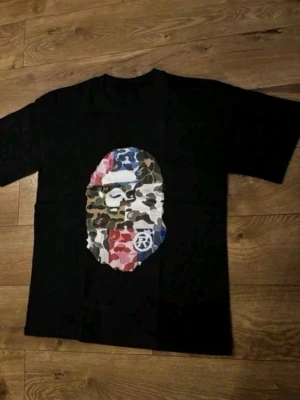 Svart bape t-shirt med kamouflage-tryck - Svart t-shirt med stort tryck av en maskerad apa i olika kamouflagefärger på bröstet. T-shirten har rund hals och korta ärmar. Perfekt för dig som gillar streetwear och unika prints.
