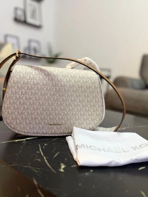 Beige axelväska från Michael Kors - Snygg beige axelväska från Michael Kors med klassiskt MK-mönster över hela väskan. Väskan har en rundad form, justerbar axelrem i brunt läder och guldfärgade detaljer. Tillverkad i tåligt material och levereras med dustbag.