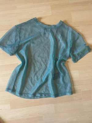 Blå top - Säljer en transparent mesh t-shirt i ljusblå färg. T-shirten har korta ärmar och är tillverkad i ett tunt, syntetiskt material som ger en cool och edgy vibe. Perfekt att styla lager på lager för en trendig look.