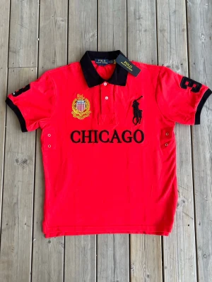 Chief Keef ”Chicago” pikétröja från Ralph Lauren - Snygg röd t-shirt från Polo Ralph Lauren med svart krage och detaljer. Stor broderad logga på bröstet, texten CHICAGO och gul brodyr på ena sidan. Svarta detaljer på ärmar och nummer 3 på höger ärm. Perfekt för dig som gillar sportig stil. Prislapp är kvar, samt qr kod. Aldrig använd.