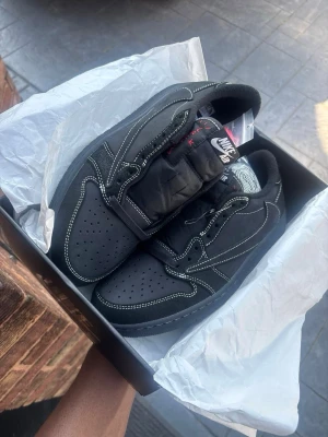 Air Jordan 1 Low Black Phantom (Travis Scott-stil) – Svart – Strl - Säljer ett par Nike Air Jordan 1 Low sneakers i helsvart med kontrasterande vita sömmar och omvänd Swoosh-logga. Skorna har en klassisk låg siluett, perforerad tåbox och detaljer i mocka och läder. Perfekt för dig som gillar streetwear och vill sticka ut.
