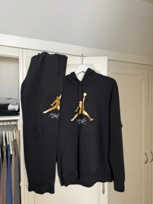 Svart Jordan set med guldlogga - Säljer ett svart set från Jordan med matchande hoodie och byxor. Båda plaggen har den ikoniska Jumpman-loggan i guld framtill och vita detaljer. Hoodien har känguruficka och dragsko i huvan. Perfekt för dig som gillar sportig och avslappnad stil.