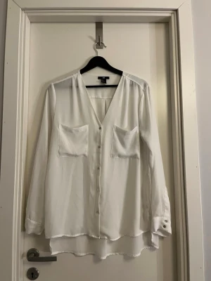 Vit blus med fickor från H&M - Stilren vit blus från H&M med v-ringning och två bröstfickor. Blusen har knäppning framtill, långa ärmar med knappdetalj vid manschetten och något längre bakstycke. Perfekt till både jeans och kjol för en trendig look.