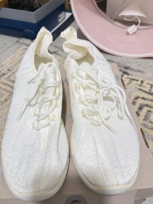 Vita sneakers med mönstrad ovandel - Säljer ett par vita sneakers med snyggt mönstrad ovandel och vita snören. Skorna har en låg profil och dragflik bak för enkel påtagning. Materialet ser ut att vara en mix av textil och syntet, vilket ger en modern och fräsch look.