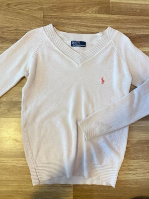 Ljusrosa stickad tröja Polo Ralph Lauren - Ljusrosa stickad tröja från Polo Ralph Lauren med V-ringning och lång ärm. Tröjan har ribbstickade muddar och en broderad rosa logga på bröstet. Perfekt för en stilren och klassisk look.