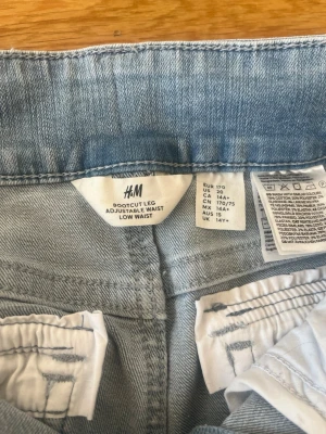 Bootcut jeans ljusblå från H&M - Ljusblå bootcut jeans från H&M med låg midja och justerbar midja. Jeansen har två bakfickor med lock och knapp, klassisk femficksdesign och är tillverkade i denim. Perfekta för en avslappnad och trendig look. Rensar garderoben och därför säljer ja dem