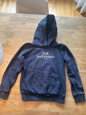 Mörkblå hoodie Peak Performance - Mörkblå hoodie från Peak Performance med stor huva och magficka. Klassisk logga i vitt på bröstet och ribbade muddar vid ärmar och midja. Barnstorlek 140 och är i fint skick!