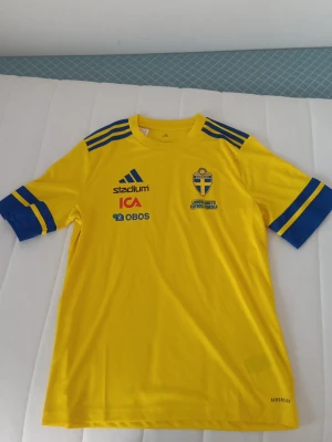 Svensk landslagströja Adidas gul - Gul svensk landslagströja från Adidas med blå detaljer och tryck. Tröjan har korta ärmar, blå ränder på axlarna och loggor från sponsorer. Materialet är lätt och andas, AEROREADY-teknologi för sportaktiviteter. Perfekt för fotbollsträning eller match.