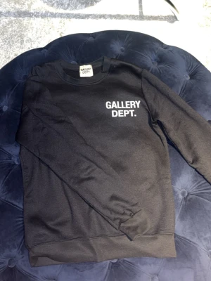Svart sweatshirt från Gallery Dept. - Cool svart sweatshirt från Gallery Dept. med vit logga tryckt på bröstet. Klassisk rund halsringning och ribbade muddar vid ärmslut och nederkant. Perfekt för en avslappnad och trendig stil.