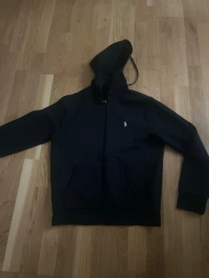 Svart hoodie med dragkedja - Svart Ralph lauren  hoodie med dragkedja och huva. Klassisk design med två fickor framtill och en liten vit logga på bröstet. Perfekt för en avslappnad stil och enkel att matcha med andra plagg.
