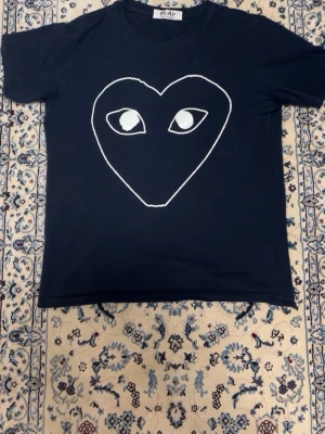 Svart t-shirt med hjärta och ögon - Säljer en svart t-shirt med ett stort vitt hjärta med ögon tryckt på bröstet. T-shirten har rund hals och korta ärmar. Perfekt för dig som gillar grafiska tryck och vill sticka ut lite extra.Tvättlappen är avklippt