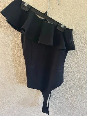 Svart one shoulder body med volang - Snygg svart body med one shoulder-design och bred volang längs urringningen. Knäppning i grenen för enkel på- och avtagning. Perfekt för dig som vill ha en trendig och stilren look. Materialet är stretchigt och bekvämt.