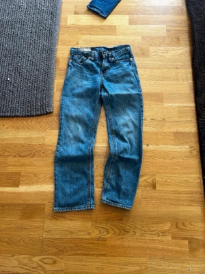 Blå raka jeans med hög midja - Säljer ett par klassiska blå jeans med rak passform och hög midja. Jeansen har fem fickor, bälteshällor och stängs med dragkedja och knapp. Materialet är kraftig denim i 100% bomull. Perfekta till en avslappnad stil.