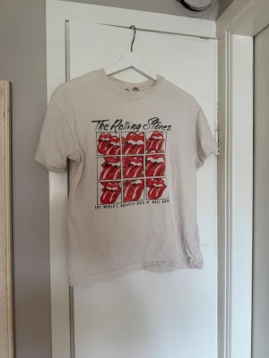 Rolling Stones vit t-shirt med tryck - Snygg vit t-shirt med klassiskt Rolling Stones-tryck framtill i rött och svart. Motiv med flera röda tungor och texten 'The Rolling Stones' samt 'The World's Greatest Rock N' Roll Band'. Perfekt för dig som gillar bandtröjor och rockstil. Lite slitningar på trycket som syns på bild.