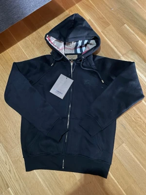 Svart hoodie från Burberry med dragkedja - Säljer en svart hoodie från Burberry med dragkedja och klassiskt rutigt foder i huvan. Hoodien har snörning i huvan, ribbade muddar och fickor framtill. Perfekt för dig som vill ha en stilren och exklusiv look. Komm med bud vid snabb affär 