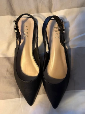 Svarta slingback ballerinaskor från Pace - Stilrena svarta ballerinaskor från Pace med spetsig tå och öppen häl. Skorna har en justerbar rem med silverfärgat spänne och är tillverkade i ett slätt, matt material. Perfekta för dig som gillar en klassisk men trendig look.