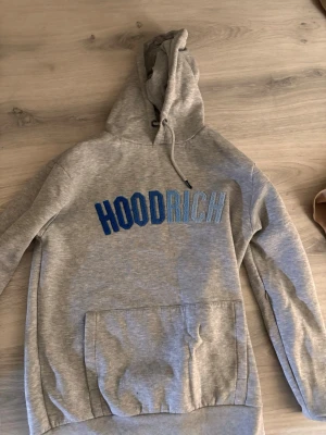 Grå HOODRICH hoodie med blå text - Snygg grå hoodie från HOODRICH med stor blå och ljusblå broderad text på bröstet. Klassisk känguruficka framtill och justerbar huva med dragsko. Perfekt för en chill och avslappnad stil.