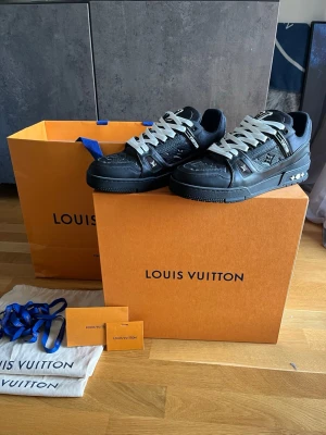 Louis Vuitton svarta sneakers - Snygga svarta Louis Vuitton sneakers med vita skosnören och diskreta monogramdetaljer på sidan och tån. Skorna har en chunky sula och lågt skaft, samt detaljer i både läder och textil. Perfekta för dig som vill ha en exklusiv och trendig look.