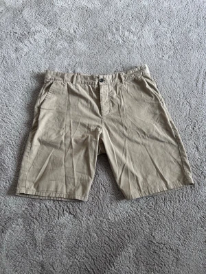 Beigea chinoshorts med fickor - Snygga beiges chinoshorts i bomull med klassisk passform. De har två sidofickor fram och två bakfickor med knappdetalj. Perfekta för varma dagar och enkla att matcha med olika stilar.