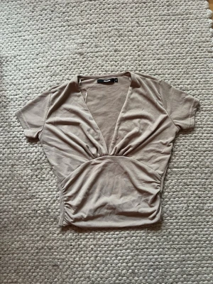 Beige topp med v-ringning - Trendig beige topp med djup v-ringning och korta ärmar. Toppen har en figurnära passform och snygga rynkdetaljer i midjan. Storlek S