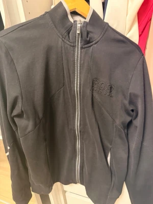 Svart zip hoodie från BOSS - Snygg svart hoodie från BOSS med dragkedja framtill och diskret broderad logga på bröstet. Hoodien har hög krage och är tillverkad i mjukt material som känns skönt mot huden. Perfekt för en clean och stilren look.