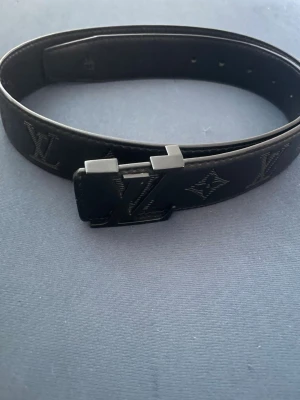 Svart  skärp i skinn - Snyggt svart skärp  i skinn med klassiskt mönster och en stilren svart spänne. Perfekt accessoar för att lyfta din outfit med en lyxig touch. Skärpet har diskreta logodetaljer längs hela bandet.