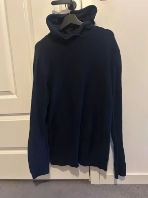 Mörkblå stickad hoodie - Mörkblå stickad hoodie med lång ärm och stor huva. Tröjan har en enkel och stilren design utan synliga detaljer eller mönster. Skriv till mig om funderingar och pris!!