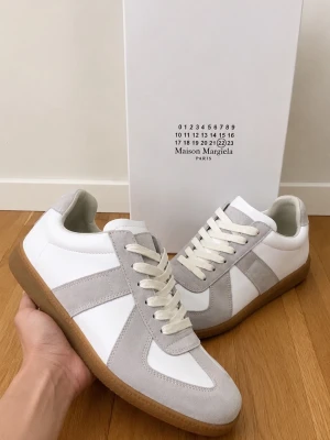 Maison Margiela vita  - Maison Margiela sneakers i vitt och ljusgrått med ovandel i läder och mocka, klassisk låg siluett och beige gummisula. Skorna har vita snören och diskret logotyp på plösen. Perfekta för en clean och stilren look.
