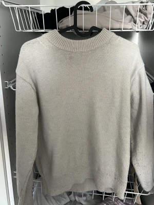 Beige stickad tröja med rund hals - En enkel och stilren beige stickad tröja med rund halsringning och långa ärmar. Tröjan har ribbade muddar vid hals, ärmslut och nederkant. Perfekt för lager på lager och passar till det mesta i garderoben.