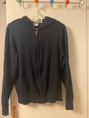 Mörkblå stickad hoodie med dragkedja - Mörkblå stickad hoodie med dragkedja framtill och långa ärmar. Tröjan har en enkel design med huva och ribbade muddar vid ärmslut och nederkant. Perfekt för en avslappnad och stilren look.