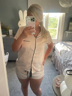 Beige kort pyjamas med svarta detaljer - Säljer en beige kortärmad pyjamas med svarta kontrastdetaljer och knappstängning framtill. Setet består av en skjorta med bröstficka och matchande shorts med svart dragsko. Perfekt för varma nätter och riktigt skön att chilla i.