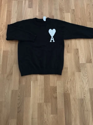 Svart stickad tröja från Ami Paris - Säljer en svart stickad tröja från Ami Paris med ett vitt hjärta och bokstaven A broderat på bröstet. Tröjan har rund hals, långa ärmar och ribbade muddar vid ärmslut och nederkant. Perfekt för dig som gillar stilrena detaljer och streetwear vibes.