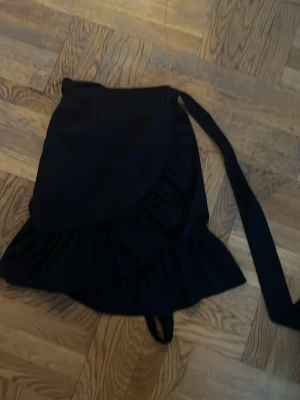 Svart omlottkjol med volang - Snygg svart omlottkjol med volangkant och knytband i midjan. Kjolen har en feminin siluett och passar perfekt till en trendig vardagslook. Materialet känns mjukt och följsamt.