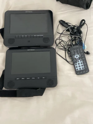 Bärbara DVD-spelare DMTECH med fjärrkontroll - Två svarta bärbara DVD-spelare från DMTECH med tillhörande fjärrkontroll och kablar. Skärmarna har inbyggda högtalare och knappar för volym, meny och uppspelning. Perfekt för resor eller bilen, enkel att använda och koppla ihop.